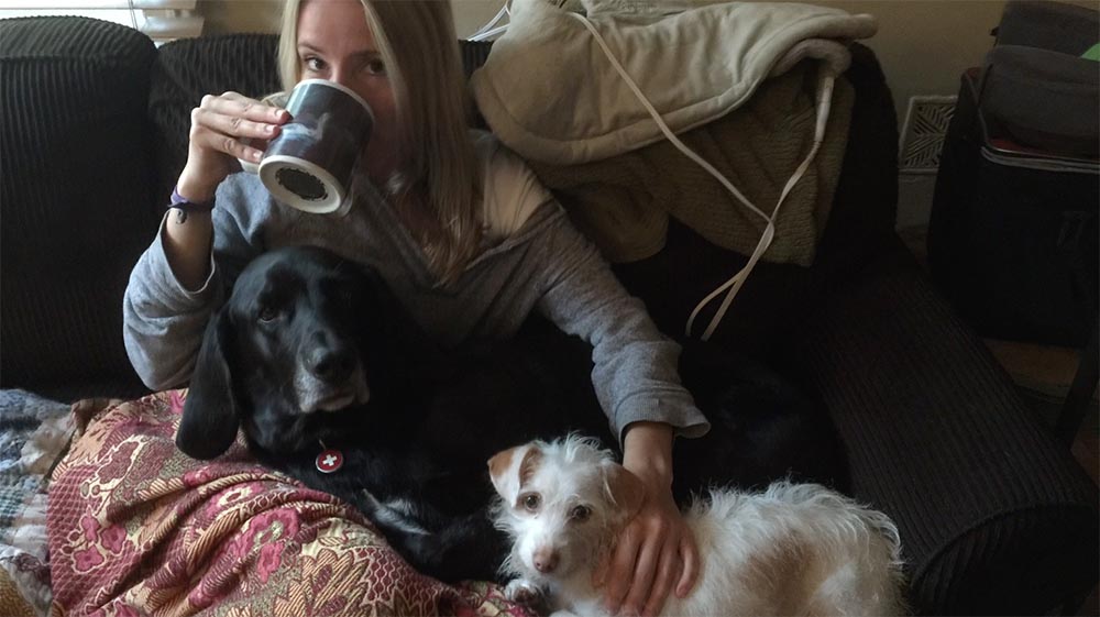 woman with grey shirt drinking coffee and relaxing on couch with 2 adult dogs