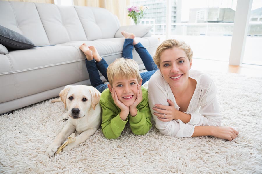 A woman in a white shirt laying on the living room white fluffy floor rug next to a boy with a green shirt and hands on face next to a white dog