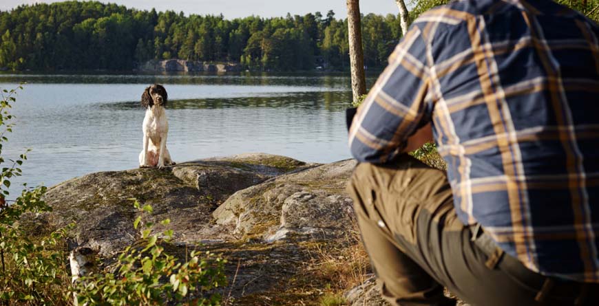 a white and black dog sitting on a rock facing a man from a distance wearing brown pants and a blue and brown plaid shirt next to a lake with green trees surrounding
